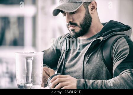 homme actif fort barbu dans la casquette et le sweat à capuche faisant des boissons à shake de protéine après une séance d'entraînement physique lourde Banque D'Images