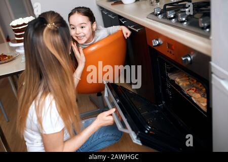 La petite fille qui attend la préparation de délicieux cookies, une jeune fille gaie assise sur la chaise et regardant sa mère. Gros plan photo Banque D'Images