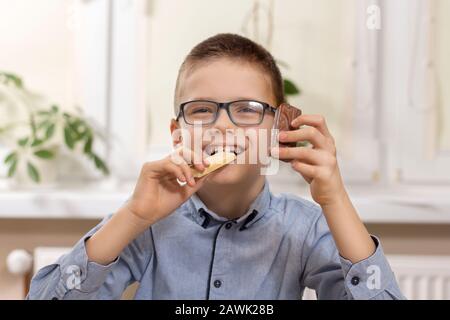 Un garçon souriant avec des lunettes se trouve à la table et tient des bonbons dans ses mains. D'une part, il tient du chocolat blanc qu'il morde et de l'autre il tient Banque D'Images