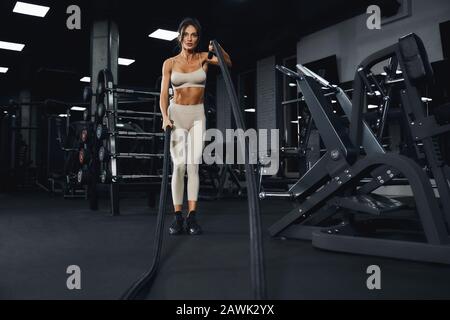 D'en dessous de la vue de la jeune femme brunette dans l'entraînement de vêtements de sport beige avec des cordes. Portrait complet de la femme de sport attrayante avec un visage fort faisant cardio dans une salle de gym vide. Concept de sport. Banque D'Images