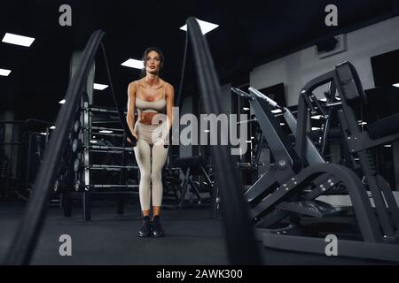 Vue de face de la jeune femme concentrée dans des vêtements de sport beige entraînement dans une salle de gym vide avec cordes. Portrait pleine longueur de fitnesswoman attrayant avec un visage fort faisant cardio. Concept de sport. Banque D'Images