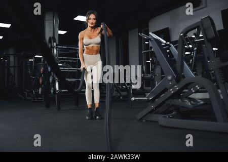 D'en dessous de la vue de la jeune femme brunette dans les vêtements de sport beige s'entraîner dans une salle de gym vide avec des cordes. Portrait pleine longueur de fitnesswoman attrayant avec un visage fort faisant cardio. Concept de sport. Banque D'Images