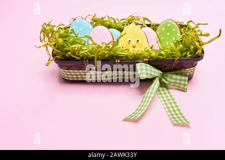 De la vue ci-dessus des cookies glacés en forme de joli poussin jaune et des œufs colorés couchés dans une boîte avec de fausses herbes vertes et un arc isolé sur fond rose. Concept de vacances de printemps et de pâques. Banque D'Images