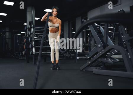 De la vue ci-dessous de la femme de la coupe brunette dans l'entraînement de vêtements de sport beige avec des cordes dans la salle de gym vide. Portrait pleine longueur de fitnesswoman attrayant avec un visage fort faisant cardio. Concept de sport. Banque D'Images