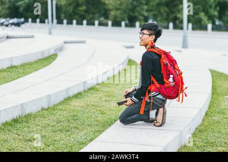 Moscou, Russie - 7 juillet 2017. Un jeune homme asiatique heureux avec un sac à dos lumineux sur ses épaules est assis sur l'escalier et le sourire. Vue latérale Banque D'Images