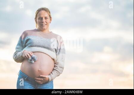 Femme enceinte posant dehors tenant de petites chaussures de bébé sur son ventre . Banque D'Images