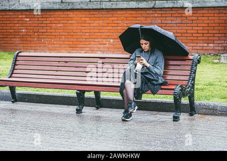 Moscou, Russie - 7 JUILLET 2017. Une femme seule dans une jupe noire et une veste grise sous un parapluie noir se trouve sur un banc et regarde le téléphone. Pluvieux Banque D'Images