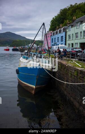 Portree, Écosse/Royaume-Uni-26 juin 2019: Les visiteurs mangent sur un banc près d'un bateau de pêche ancré dans le Loch Portree, situé près d'une maison peinte aux couleurs vives Banque D'Images