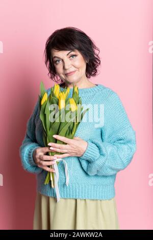 Belle vieille femme avec maquillage professionnel fumé et tenue de coiffure fleurs de tulipes sur fond rose Banque D'Images