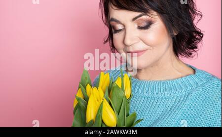 Belle vieille femme avec maquillage professionnel fumé et tenue de coiffure fleurs de tulipes sur fond rose Banque D'Images