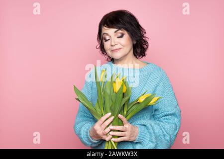 Belle vieille femme avec maquillage professionnel fumé et tenue de coiffure fleurs de tulipes sur fond rose Banque D'Images