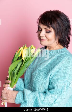 Belle vieille femme avec maquillage professionnel fumé et tenue de coiffure fleurs de tulipes sur fond rose Banque D'Images
