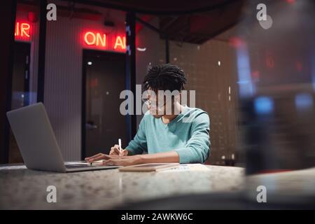 Portrait grand angle de jeune homme afro-américain travaillant tard à la maison ou dans le sombre bureau, espace de copie Banque D'Images