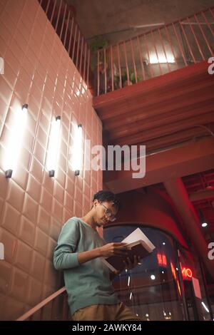 Portrait à bas angle de jeune homme afro-américain tenant des livres tout en se penchant sur le mur dans le campus de l'université, espace de copie Banque D'Images