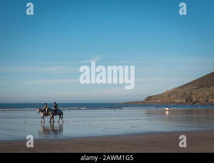 Deux chevaux sont montés le long de la plage de Saunton Sands au nord de Devon, en Angleterre, au soleil avec des surfeurs en arrière-plan Banque D'Images