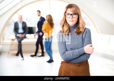 Portrait tourné d'une femme d'affaires d'âge moyen en charge au bureau pendant que les gens d'affaires consultent en arrière-plan. Banque D'Images