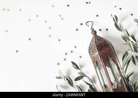 Vintage bronze marocain, lanterne arabe. Feuilles d'olive vertes, branches isolées sur fond de table blanc, étoiles dorées confetti. Carte de vœux pour Banque D'Images