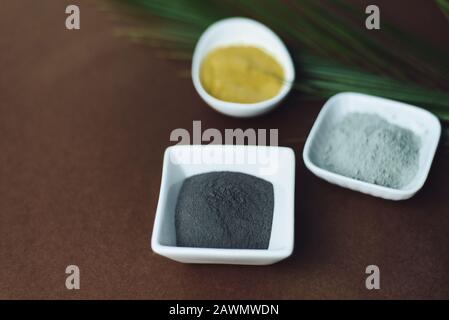 Argile noire, jaune et grise dans un bol en porcelaine blanche sur fond brun avec une feuille de palmier. Cosmétiques naturels pour soins à domicile ou en salon, spa Copy Banque D'Images
