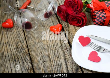 Coeurs sur une plaque avec а fourchette et couteau, boîte cadeau, corkvec, deux vitivinicoles, roses rouges et bougies sur une vieille table en bois. Mise au point sélective. Banque D'Images