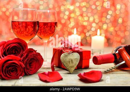 Coeur en bois, roses rouges, boîte à cadeaux, bouteille de vin, corkscrew, verres de vin et bougies brûlantes sur une vieille table en bois devant une lumière floue Banque D'Images