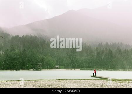 Personne imreconnaissable avec chien debout dans la tempête de pluie sur une jetée en bois d'un lac de montagne, avec un parte de pin vert luxuriant et des nuages brumeux Banque D'Images
