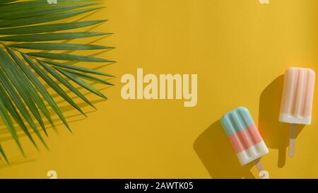 Vue de dessus de deux popsicules colorés avec feuilles de noix de coco, concept créatif minimal d'été sur fond jaune Banque D'Images