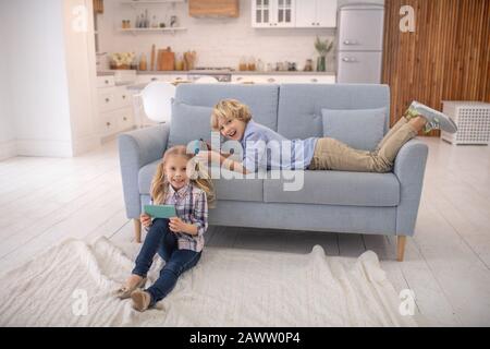 Garçon allongé sur le canapé, fille assise sur le sol avec des gadgets Banque D'Images