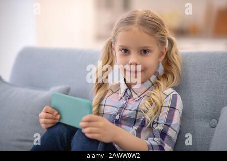 Fille assise sur canapé, tenant smartphone et souriant Banque D'Images