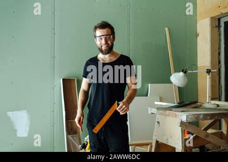 Bel homme souriant barbu debout avec une règle d'angle dans un appartement rennovant Banque D'Images