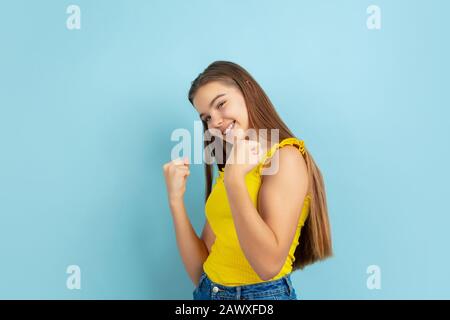 Gagnant réussi. Portrait de jeune fille caucasienne isolé sur fond bleu. Magnifique modèle en tenue jaune décontractée. Concept d'émotions humaines, expression faciale, ventes, publicité. Copyspace. C'est mignon. Banque D'Images