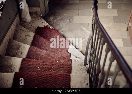 En regardant les escaliers cassés et sales avec un tapis rouge et une rampe en bois dans un bâtiment abandonné et bourré. Banque D'Images