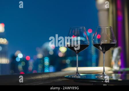 Deux verres de vin rouge sur la table du bar sur le toit avec des lumières colorées de la ville. Banque D'Images