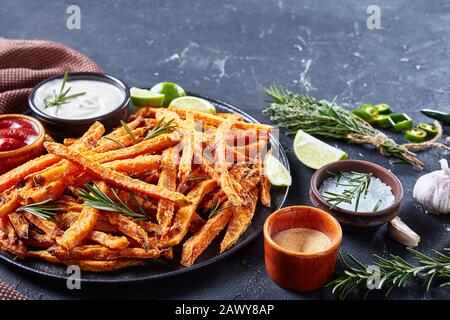 Gros plan de frites croustillantes de patates douces avec sauce et ketchup sur un plateau noir sur une table en béton avec chaux et ingrédients, vue horizontale de l'ab Banque D'Images