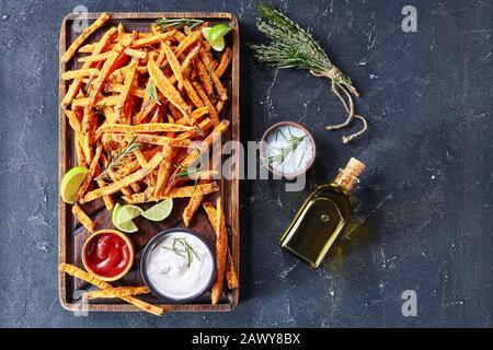 Gros plan de frites de pommes de terre salées croustillantes avec sauce et ketchup sur une planche en bois impolie sur une table en béton avec de la chaux et un bouquet d'herbes aromatiques, Banque D'Images
