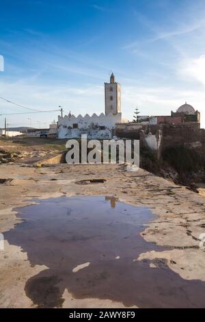 Petite mosquée construite sur un rocher sur la côte de l'océan Atlantique avec une plage de sable. Des flaques dans la rue après une pluie de nuit. Ciel bleu avec nuages. Moulay Banque D'Images