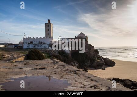 Petite mosquée construite sur un rocher sur la côte de l'océan Atlantique avec une plage de sable. Des flaques dans la rue après une pluie de nuit. Ciel bleu avec nuages. Moulay Banque D'Images