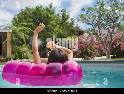 Femme appliquant, vaporisant de la crème solaire sur bras sur radeau gonflable dans la piscine ensoleillée Banque D'Images
