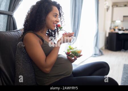 Jeune femme enceinte souriante mangeant une salade sur un canapé Banque D'Images