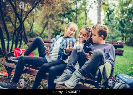 Groupe d'amis jouant à des jeux vidéo sur smartphone après l'école tout en étant assis sur le banc dans le parc. Banque D'Images