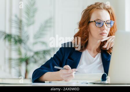 Un entraîneur féminin expérimenté et sérieux fait des records dans le bloc-notes, concentré à l'écran de l'ordinateur portable, a de longs cheveux rouges, porte des lunettes transparentes, montres Banque D'Images