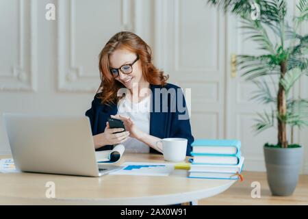 Technologie et concept de communication. La jeune femme aux cheveux rouges vérifie la boîte de courriel sur le téléphone cellulaire moderne, a un sourire agréable, étudie les documents, possède grand c Banque D'Images