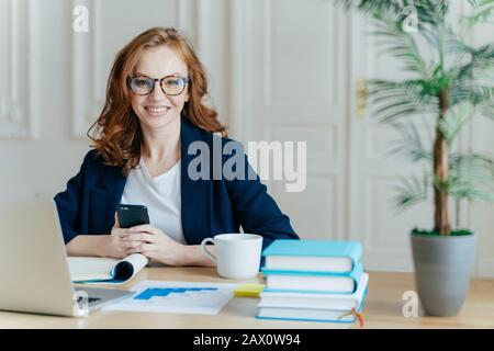 Photo de la jeune femme souriante aux cheveux rouges installe l'application sur le téléphone mobile, vérifie le flux de nouvelles, travaille sur l'ordinateur portable à distance, pose dans l'espace de travail Wit Banque D'Images