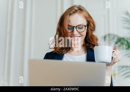 La tête de la femme souriante aux cheveux rouges dans les verres optiques, aime le café chaud ou le thé, concentré dans l'ordinateur portable, fait des achats en ligne, sourires agréables Banque D'Images