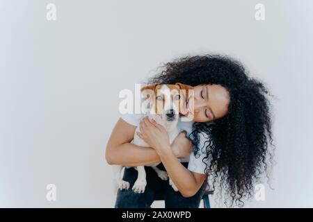 Image d'une jolie femme joueuse avec coiffure Afro, sourires gladeusement, tient animal domestique, porte chien à veterre, isolé sur fond blanc. G Banque D'Images