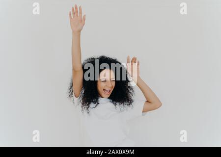 Joyeuse curieuse femme africaine américaine lève les mains, rires du bonheur, célèbre le triomphe, se réjouit du succès, porte un t-shirt blanc, pose intérieur, g Banque D'Images