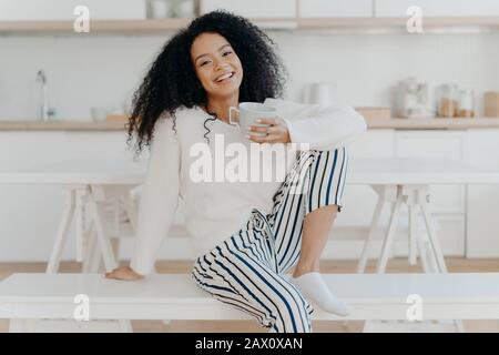 Belle jeune femme avec coupe de cheveux Afro, pose sur un banc blanc intérieur, vêtu de vêtements élégants, aime les boissons aromatiques, aime le petit déjeuner à la maison Banque D'Images