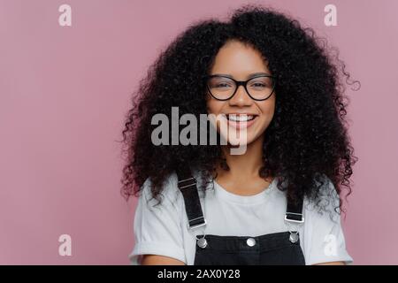 Gros portrait d'une femme à peau foncée avec des cheveux nets, sourires heureux, porte des lunettes optiques, vêtements décontractés, pose sur fond violet exprime Banque D'Images