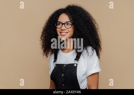 Une femme rêveuse et heureuse à la coiffure décontractée, regarde à travers des lunettes transparentes, vêtues d'un t-shirt blanc décontracté et d'un salopette noire, pose contre le brun Banque D'Images