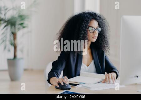 Une femme d'affaires sérieuse et curieuse, centrée sur l'affichage de l'ordinateur, travaille sur la réalisation du projet, entourée de manuels et de papiers, porte des lunettes pour le décor de vision Banque D'Images
