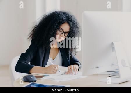Photo de sérieuse Afro femme écrit dans des papiers, assis à la table avec ordinateur moderne, article de créats dans le journal, porte des lunettes et des lunettes, pose à l'intérieur Banque D'Images